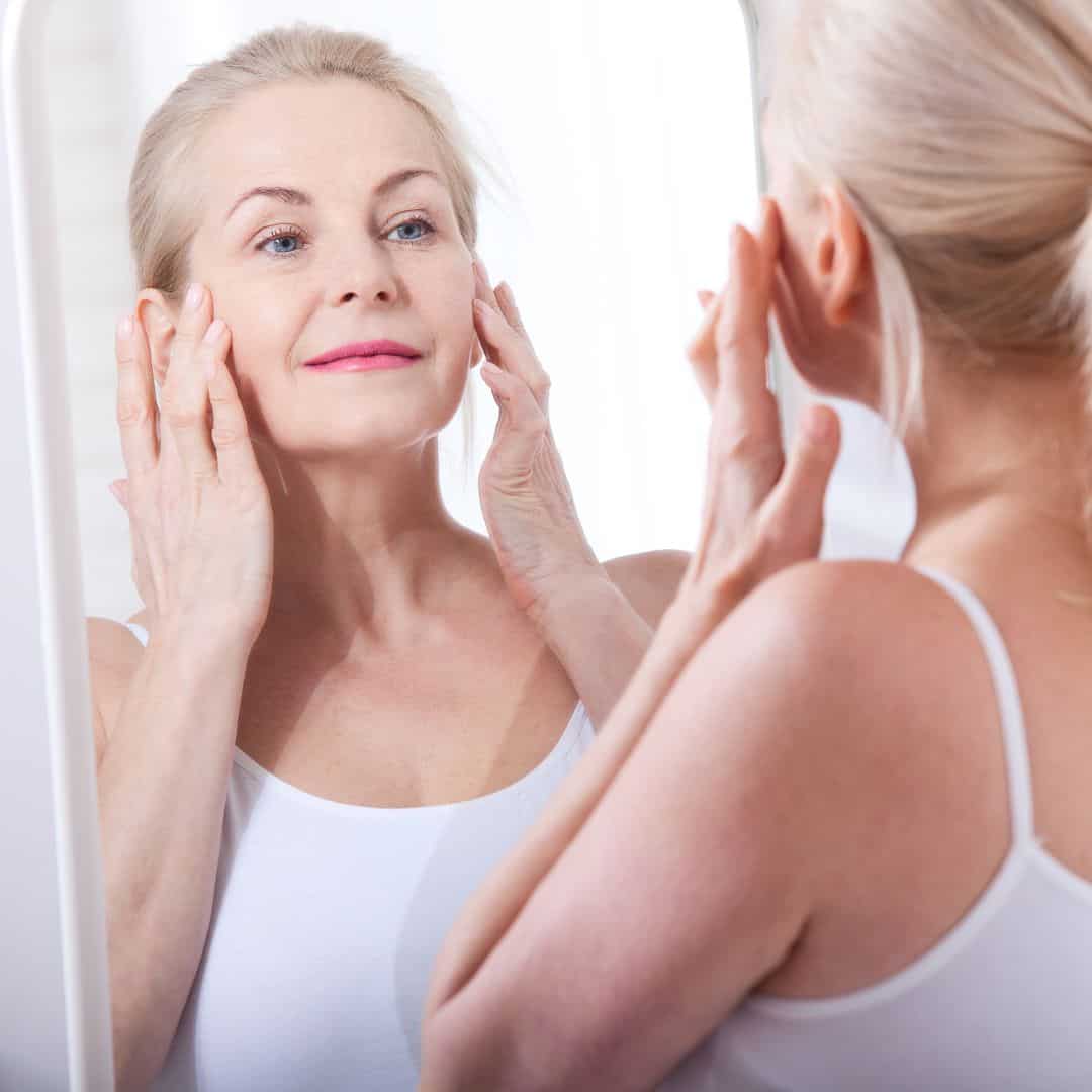 Woman examining her face in mirror for skin tightening treatment.